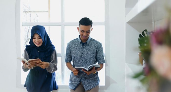 a lighter-skinned Asian woman wearing a hijab and reading, and a lighter-skinned Asian man laughing and reading