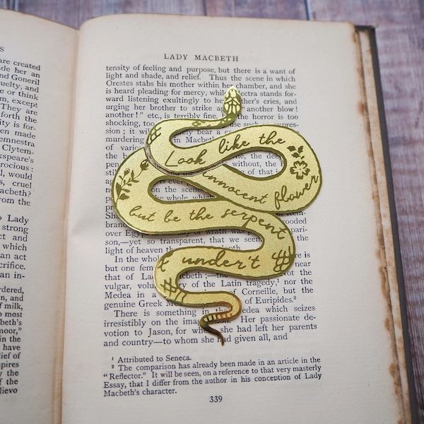 a brass bookmark in the shape of a snake. In the body of the snake are the words "look like the innocent flower but be the serpent under't"