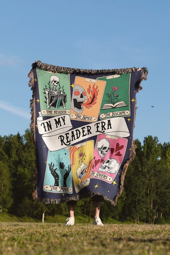 Person standing outside holding up a large fringed throw blanket that reads "In My Reader Era" surrounded by colorful tarot cards 