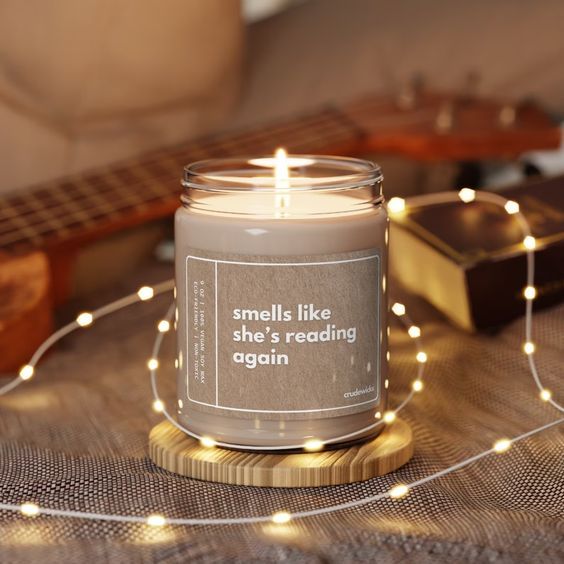 tan colored candle reading "smells like she's reading again" on a wood table surrounded by twinkle lights 