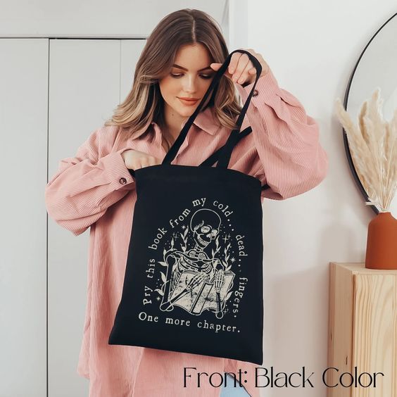 a blonde white woman holding a black tote bag with an image of a skeleton holding a book. The words say "You can pry my book from my cold, dead, fingers. Just One More Chapter."