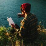 fair-skinned person readinng a book with a mug in their hand; they're on a very scenic cliff