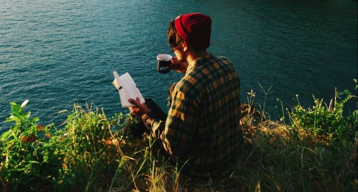 fair-skinned person readinng a book with a mug in their hand; they're on a very scenic cliff