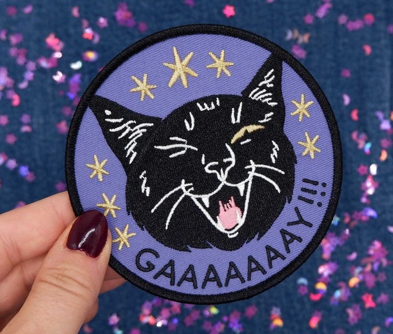 an embroidered patch of a cat winking and yowling with the word gaaay!! underneath