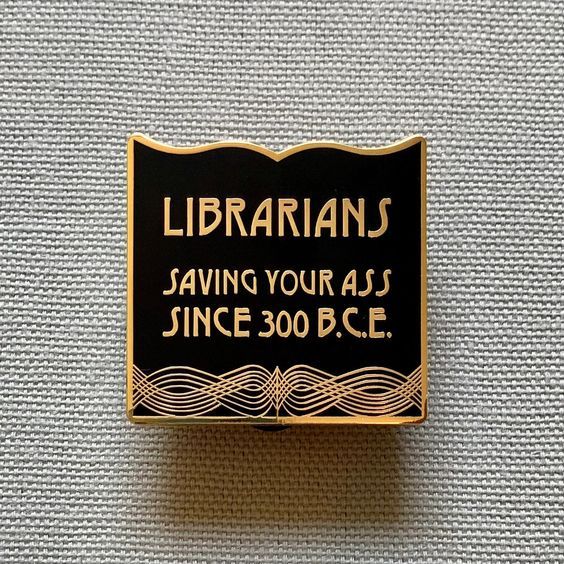 a black and gold enamel pin that says "librarians, saving your asses since 300BC"