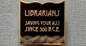 a black and gold enamel pin that says "librarians, saving your asses since 300BC"