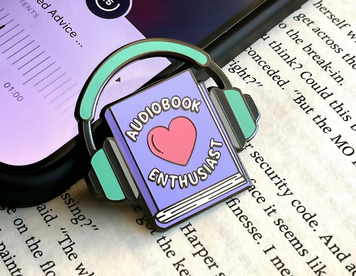 an enamel pin of a purple book wearing headphones that says "audiobook enthusiest"