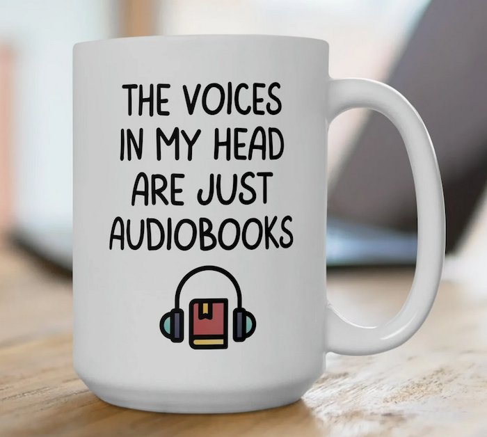 a white mug that says "the voices in my head are just audiobooks" with a graphic illustration of a book wearing headphones