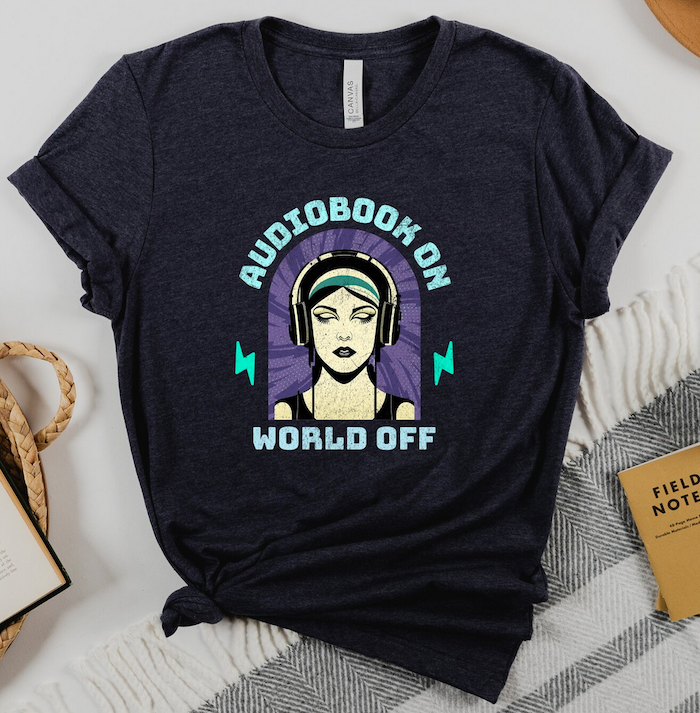 a dark grey shirt with a graphic illustration using aqua and purple of a woman wearing headphones that says "audiobook on world off"