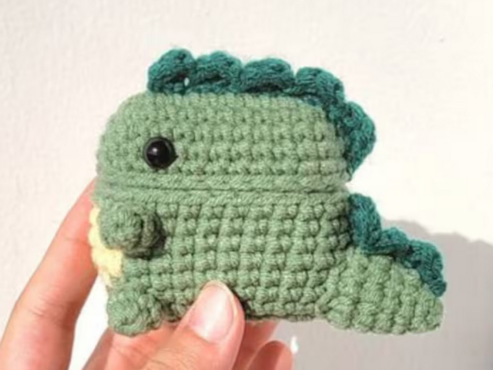 a crocheted green dinosaur to cover an airpod case on etsy by EmbroideryGiftsVN