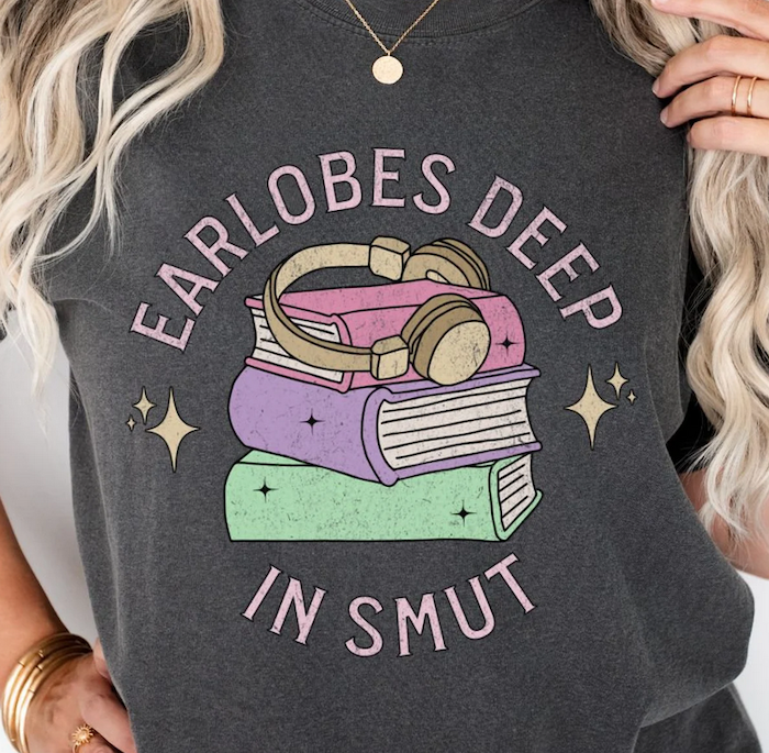 dark grey tshirt with graphic illustration of books and headphones that says "earlobes deep in smut"