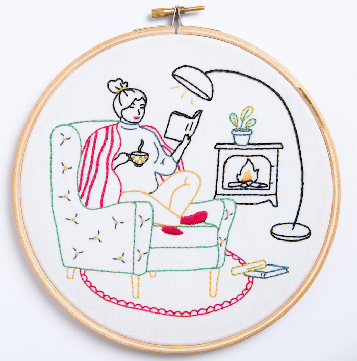 an embroidery kit that makes an image of a woman sitting in a chair holding a mug and reading a book