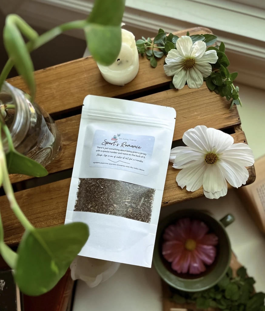Image of a bag of loose leaf tea with a label that says "Sports romance tea", displayed next to flowers and a mug