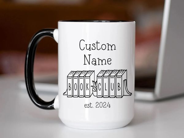 white coffee mug with a black handle. Black text on the mug reds "custom name est. 2024" is positioned above a graphic of a row of books