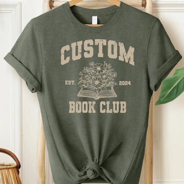 an olive green tshirt with beige text that reads "Custom Book Club" wrapped around a line drawing of an open book with flowers growing out of it 