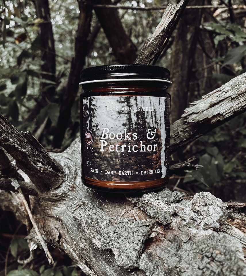 Image of a candle in a glass jar with a label that says "books and petrichor" displayed on a log