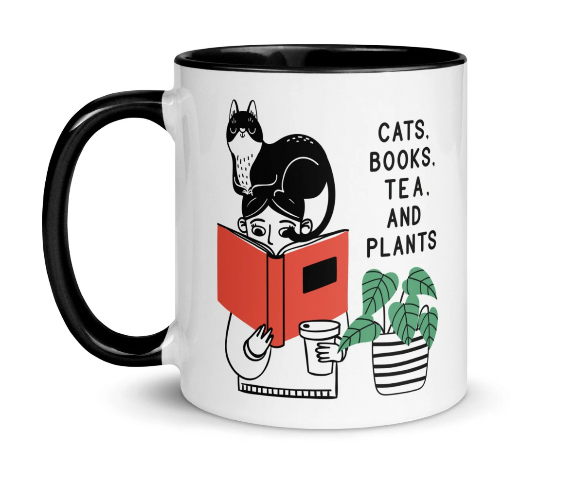 A white mug with black handle and interior. On the front of the mug is the phrase "Cats, books, tea, and plants," along with a drawing of someone reading a book with a cup of tea in hand and a cat perched on their head next to a plant