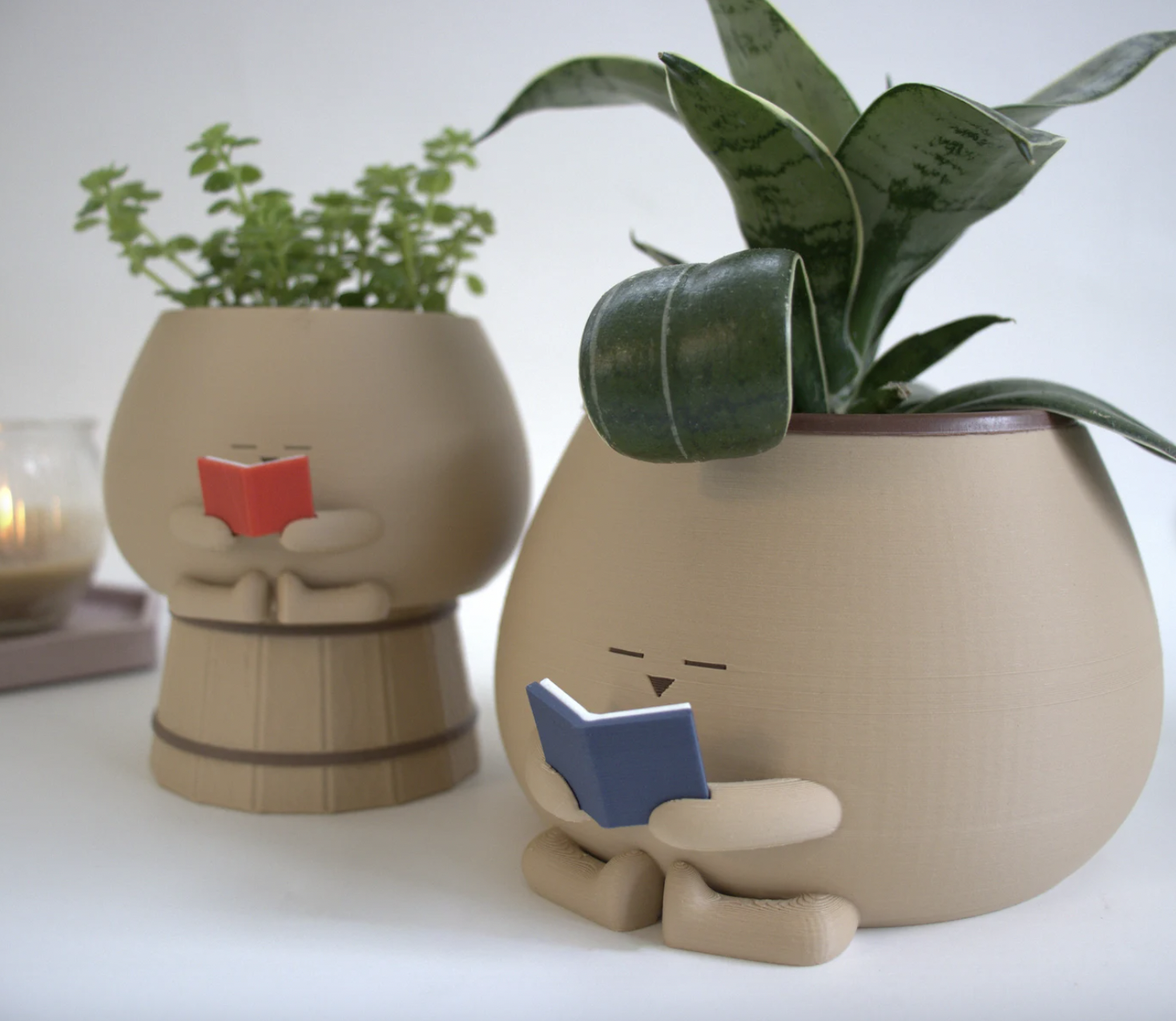 Image of two ceramic planters with tiny faces and arms holding a book. Both have greenery coming out of the top.