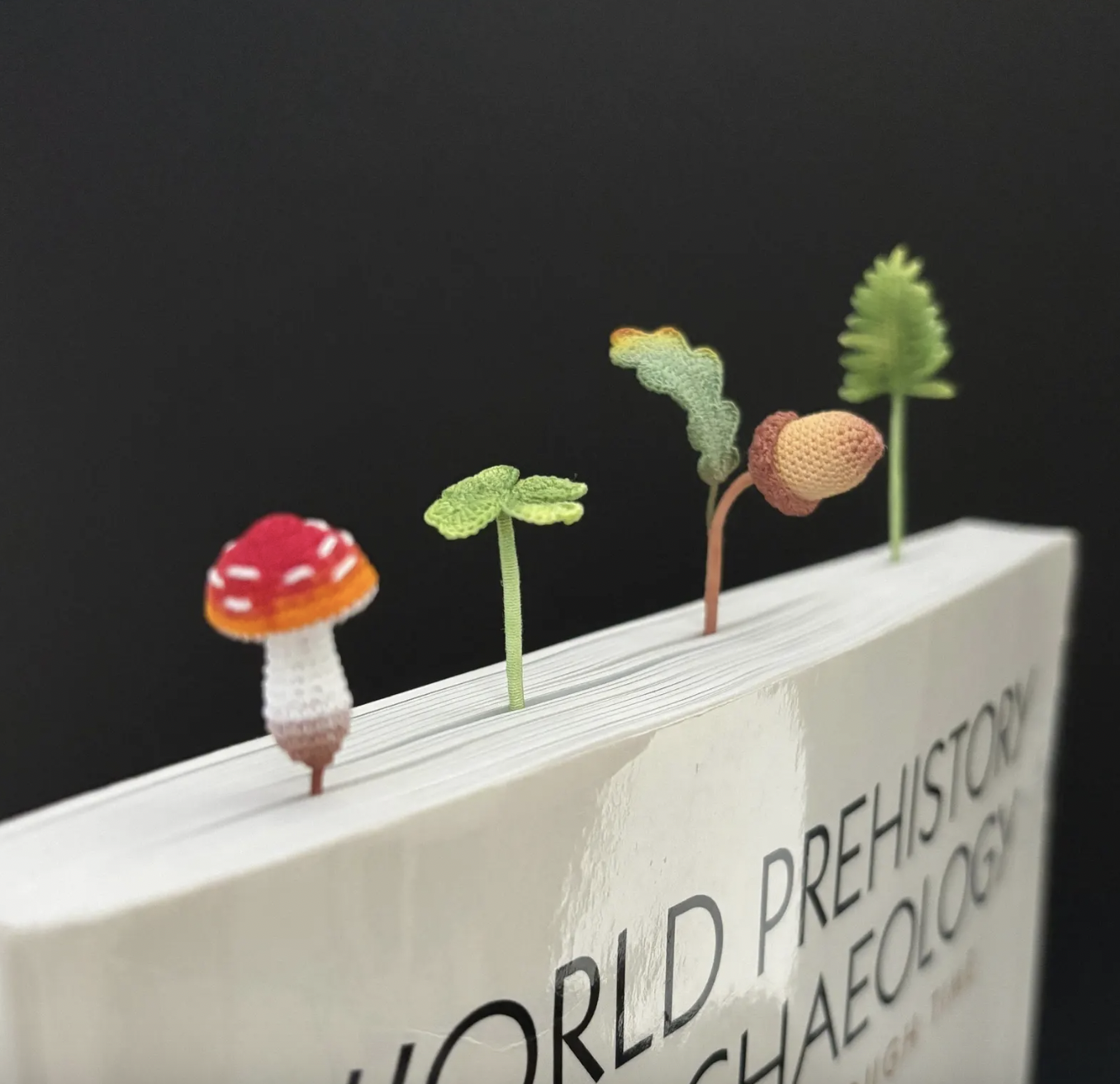 Image of tiny crocheted bookmarks poking out of the top of a book, including a mushroom, a clover, an acorn, and a fern leaf