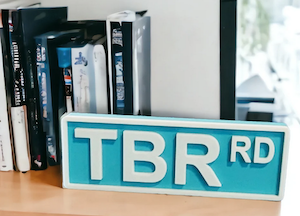 a wooden sign on a bookshelf meant to look like a street sign that says TBR RD