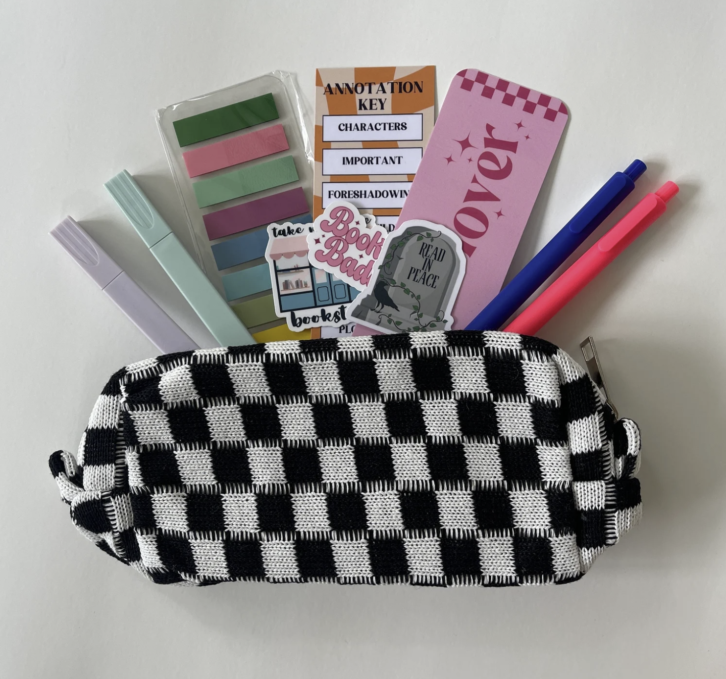 A black and white checkered pouch filled with supplies for book annotation including colored pens and highlighters, sticky note tabs, stickers, and bookmarks fanning out from inside the pouch.