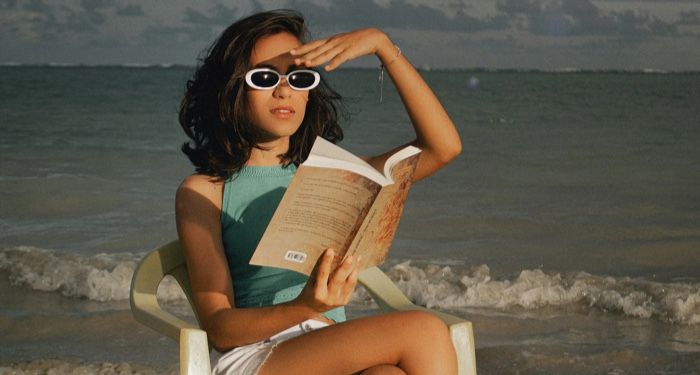 a lightly tanned-skin woman reads a book while sitting in s a white chair on the beach