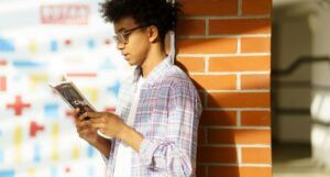 brown-skinned Black man with afro reading a book against a brick wall