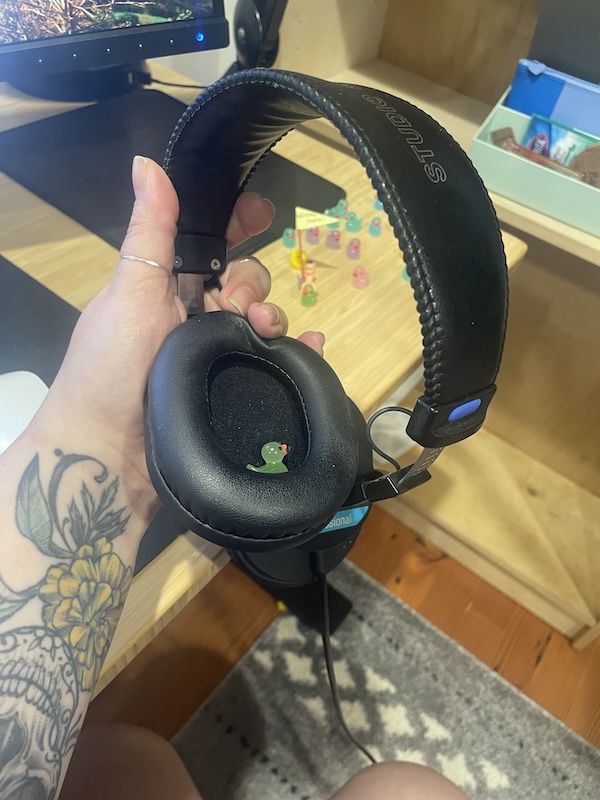 a tiny plastic neon green duck tucked inside an over-ear headphone with another handful of ducks of assorted colors on a desk in the background