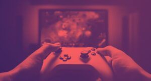 hands holding a video game controller with a TV playing a video game in the background. The image is washed in a red and back filter