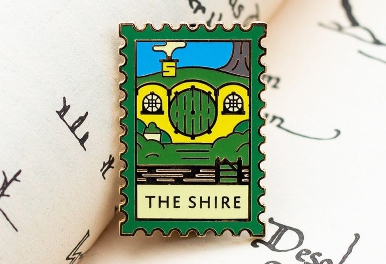 enamel pin in the style of a stamp with an illustration of a hobbit hole and the text The Shire