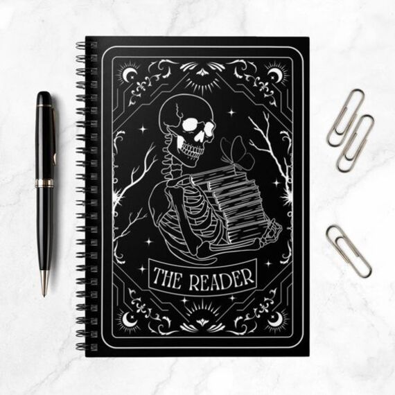 a black notebook with a tarot design on the cover of a skeleton holding books and the caption "the reader"