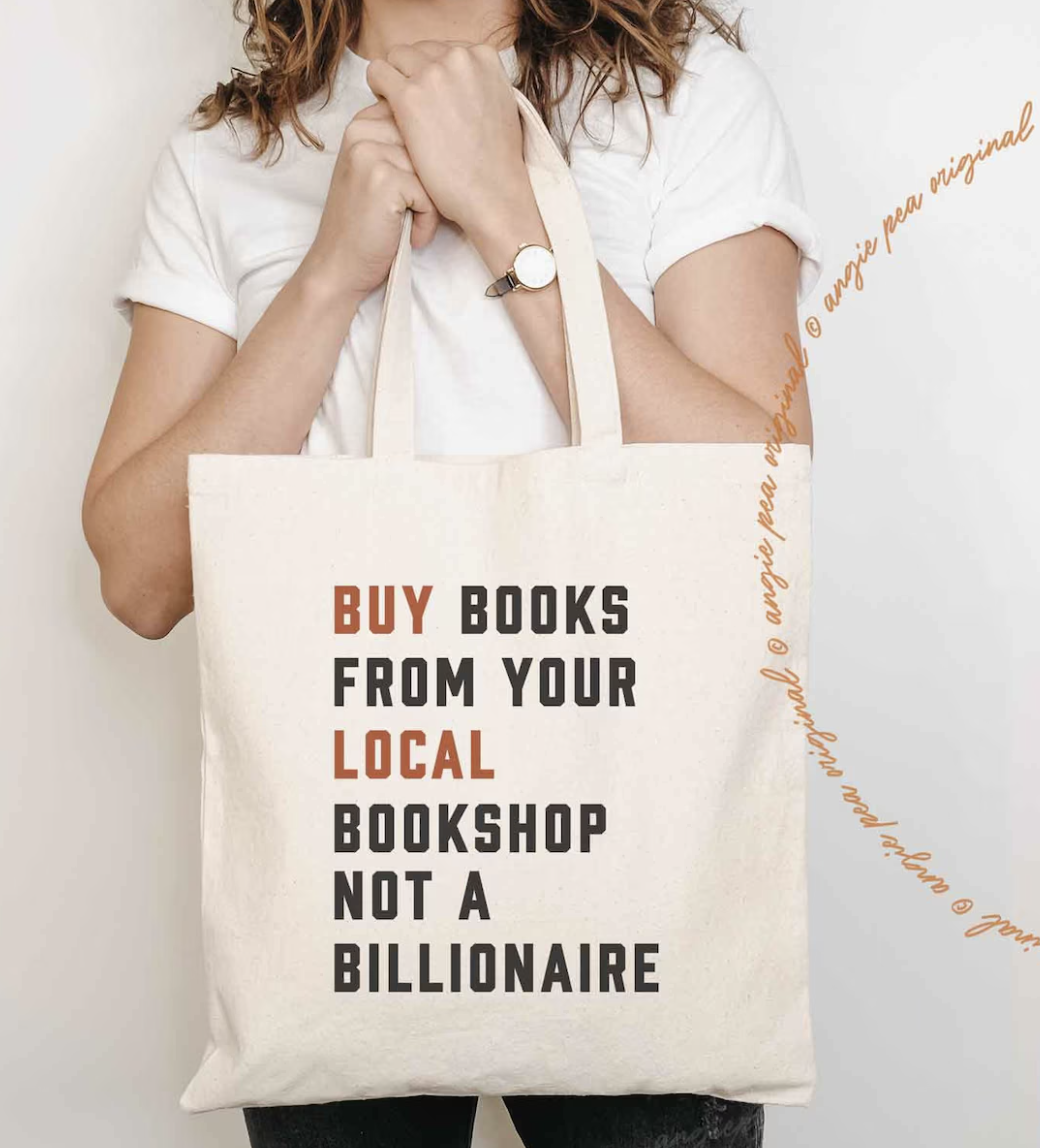 Image of someone holding a cream colored tote bag with "buy books from your local bookshop, not a billionaire" screen printed on the front