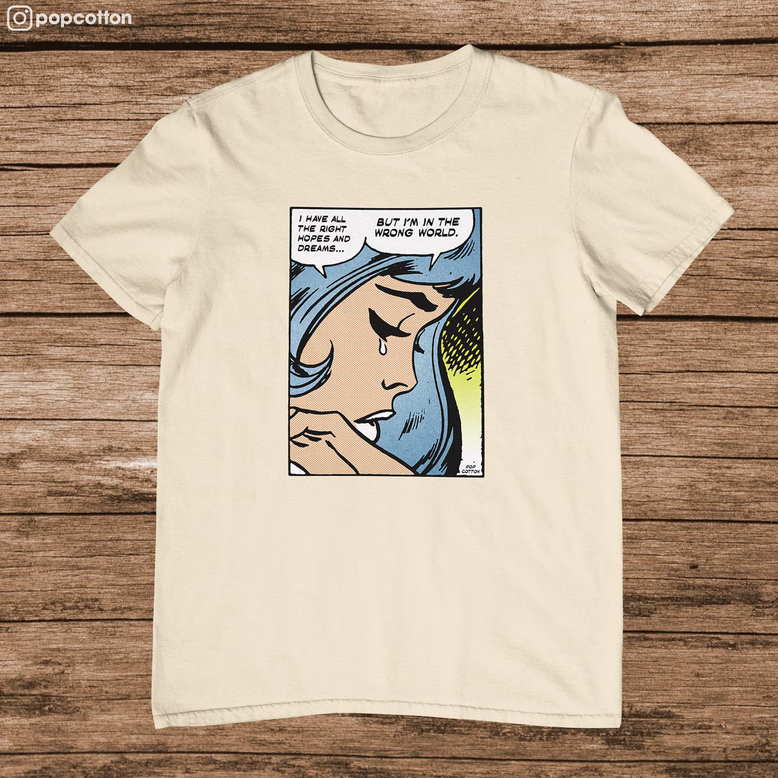 A cream colored shirt featuring a comic book panel with a blue-haired woman saying "I have all the right hopes and dreams...but I'm in the wrong world"