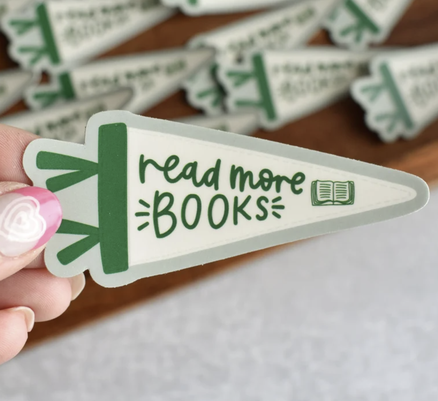 Image of a white woman's hand holding a sticker in the shape of a pennant with a green stripe that says "read more books"