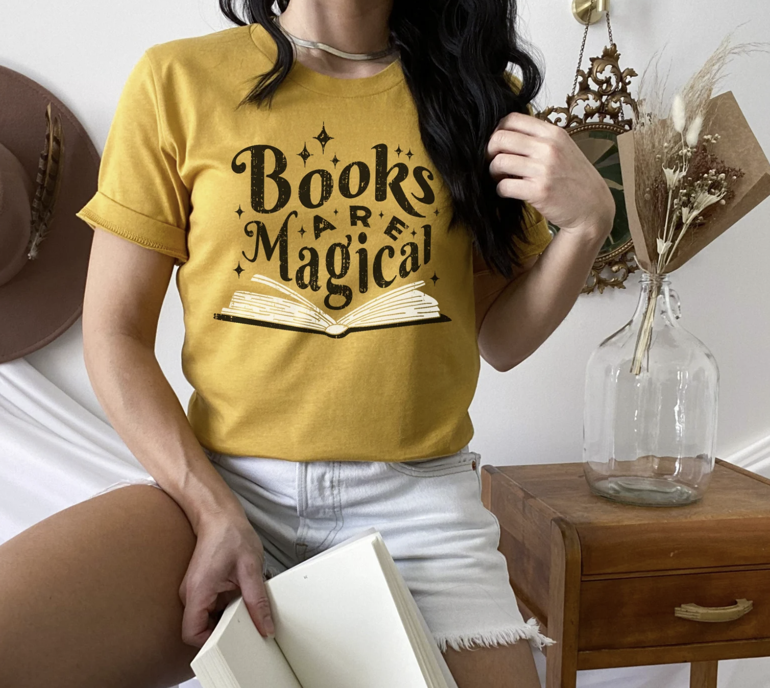 Picture of a white woman with dark hair posing with an open book in one hand and wearing a golden yellow shirt with an image of an open book with the words 