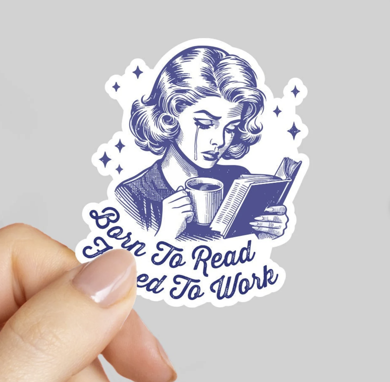 Image of a well-groomed white woman whose fingers are holding a blue and white sticker with a vintage drawing of a woman crying while reading a book and drinking coffee that says "Born to read, forced to work"