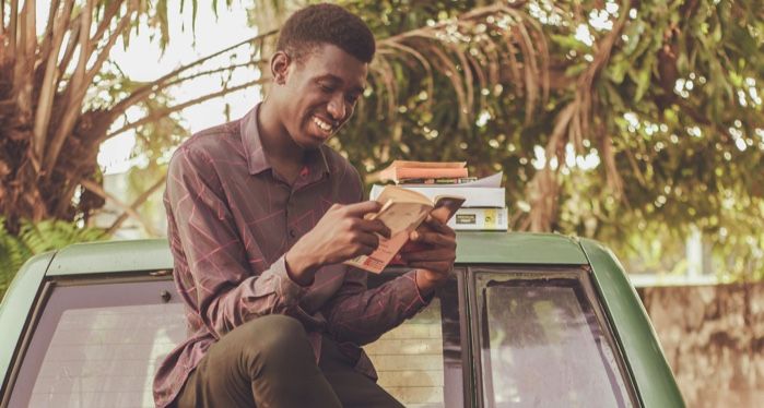 a dark brown-skinned Black man smiling and reading a book while sitting on a jeep with palm trees in the back