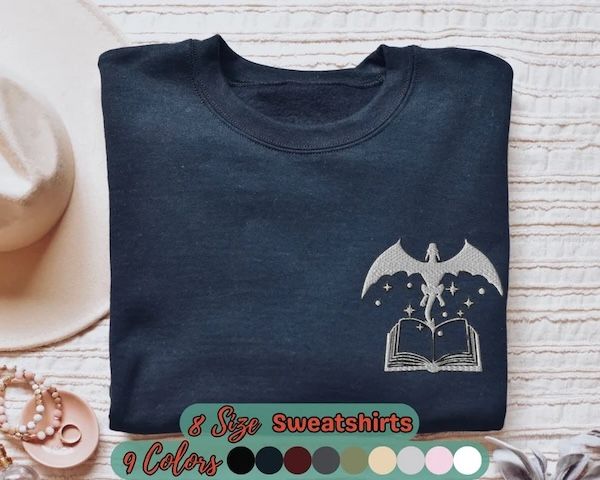 navy blue crewneck sweater with an embroidered design in the upper left of a dragon emerging from inside a book