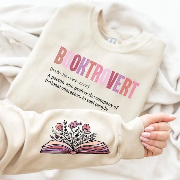 cream crewneck sweater with pastel text that reads "booktrovert." Under that is simple black text with a pronunciation guide and definition of "a person who prefers the company of fictional characters to real people" 