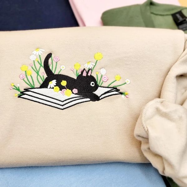 cream crewneck sweater with an embroidered design of a black cat and flowers resting inside of a book