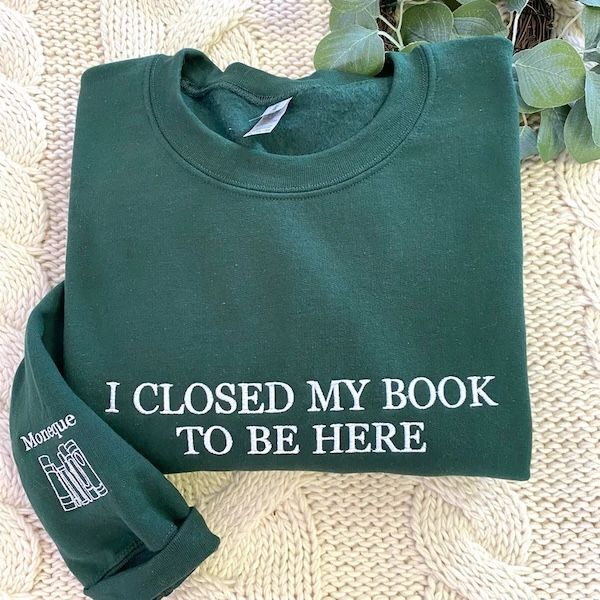 green crewneck sweater with white embroidered text that reads "I closed my book to be here"