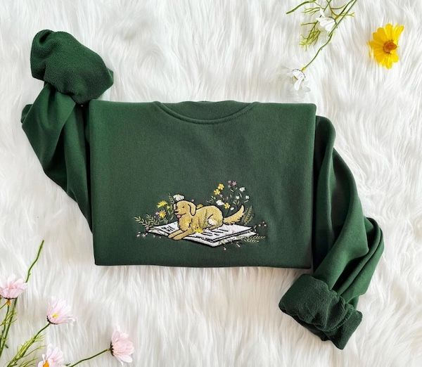 green crewneck sweater with an embroidered design of a black dog and flowers resting inside of a book