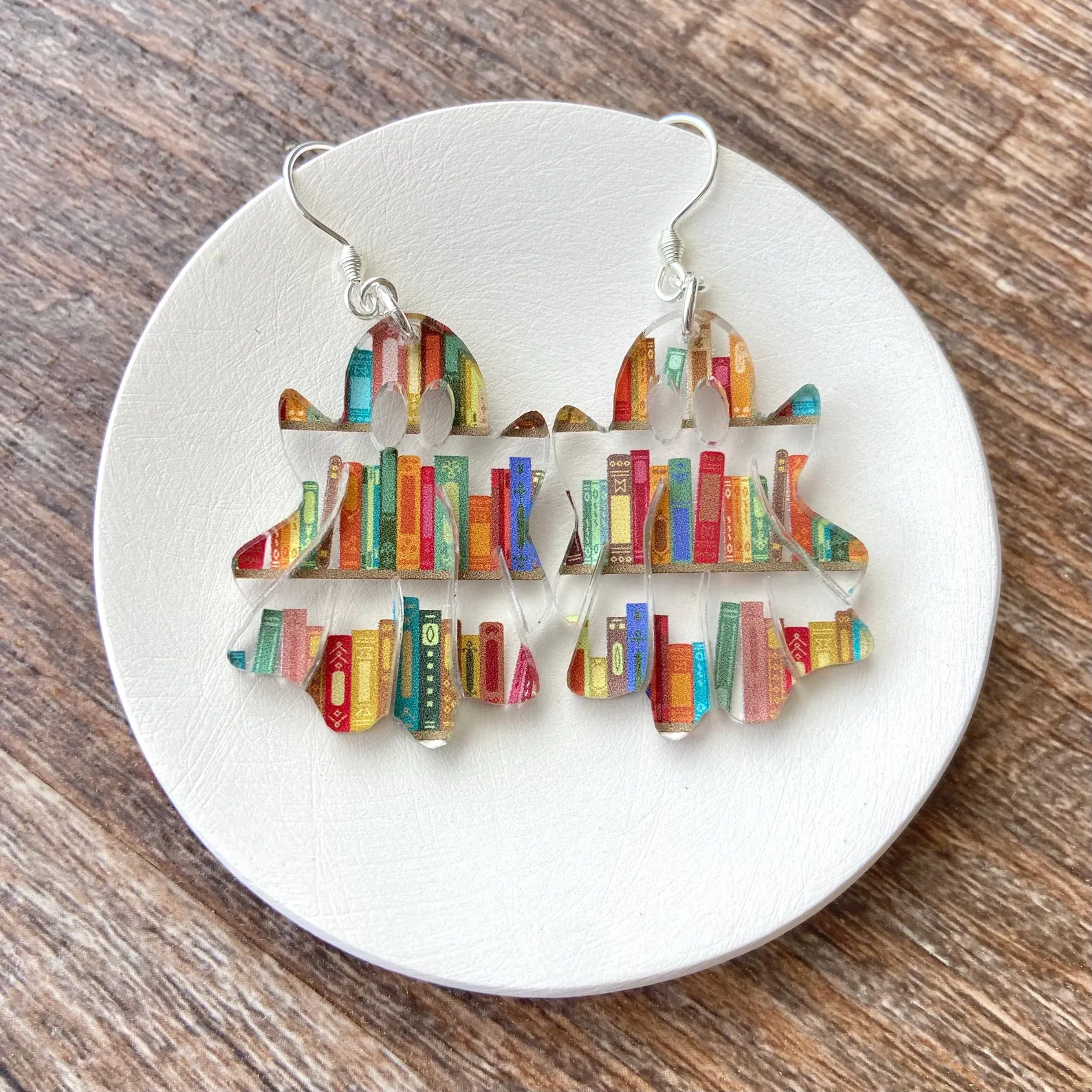 Image of a pair of clear earrings that feature books on three sets of shelves. The sets are designed to look like ghosts. 