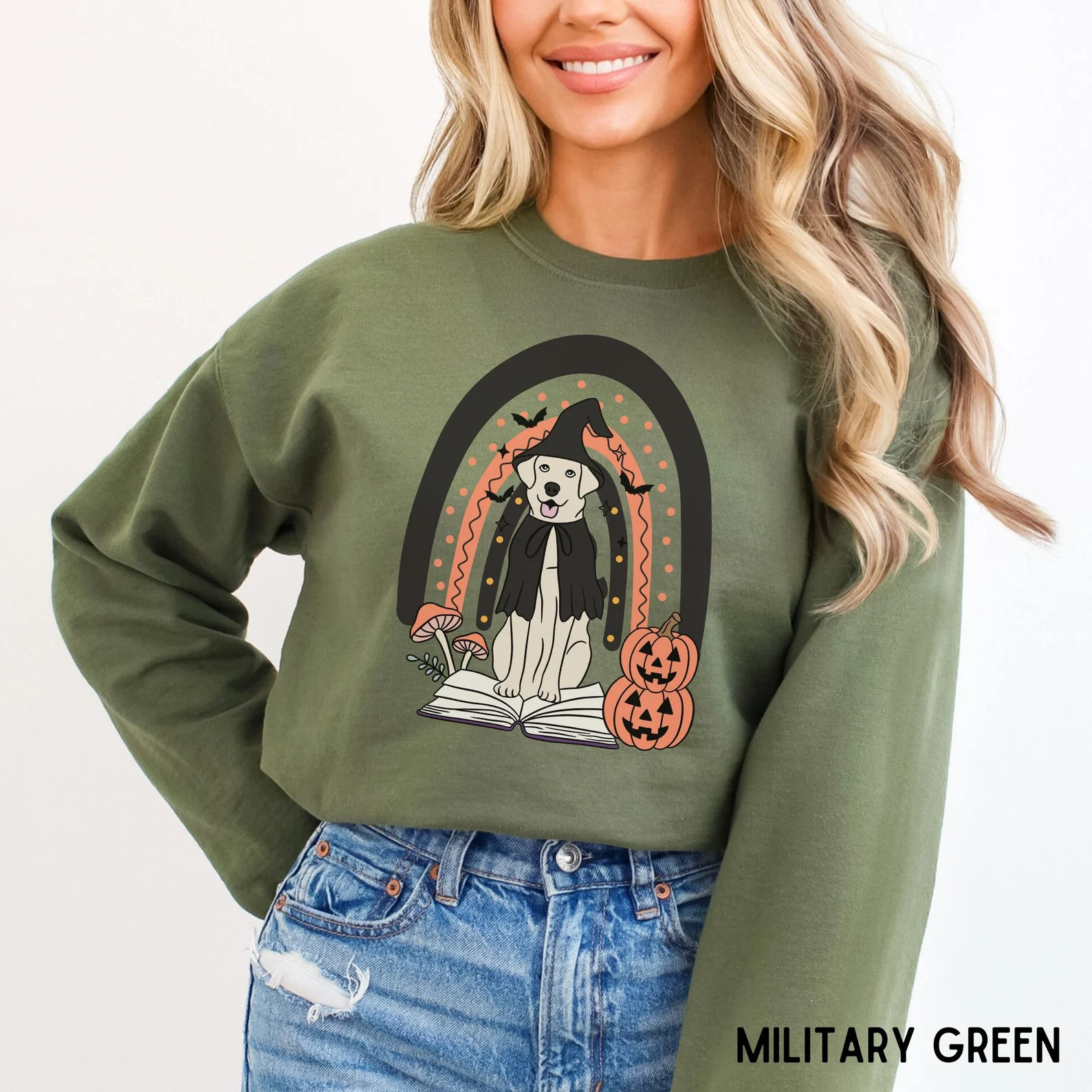 Image of a white woman in a green crewneck sweatshirt. It has a dog dressed up as a witch with an open book and pumpkins. 