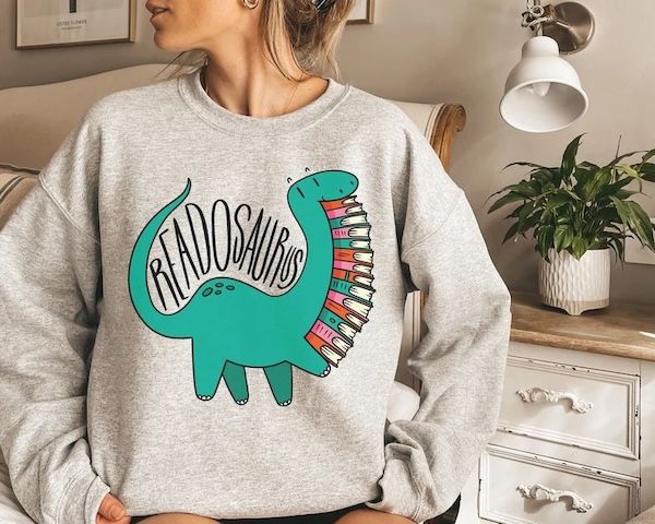 light grey crewneck seater with a large green dinosaur on the front holding a large stack of books. the word "Readosaurus" hovers above its back