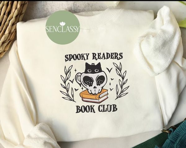 cream crewneck sweater with black text on the front that reads "Spooky Readers Book Club." the text is arranged around a graphic of a small black cat with large eyes inside of a skull mug on a stack of books