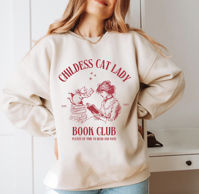 a cream colored sweatshirt with an illustration of a woman and cat reading with text saying "childless cat lady bookclub plenty of time to read and vote"