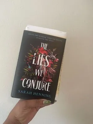a brown-skinned hand with green nails holding The Lies We Conjure by Sarah Henning
