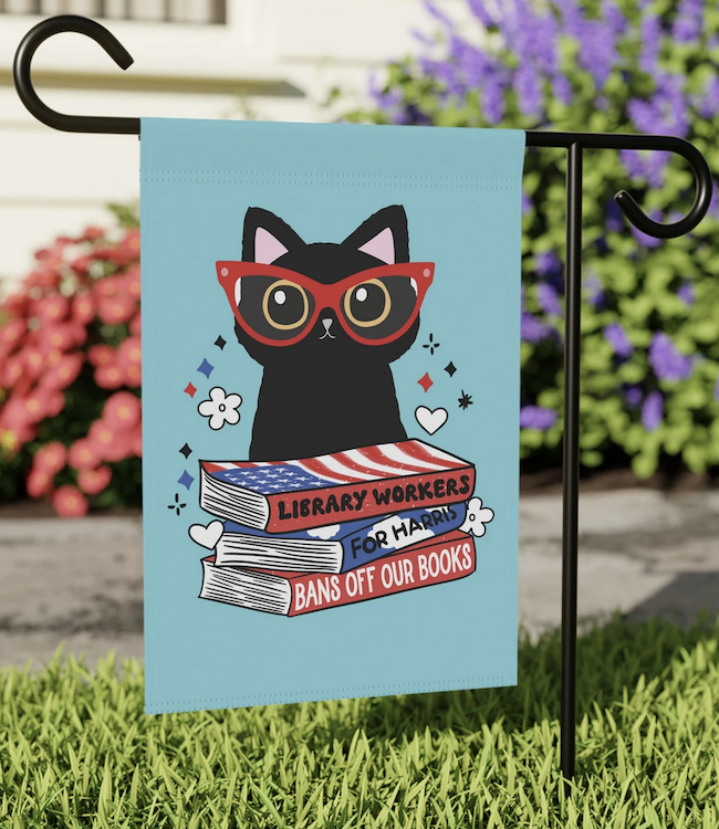 a blue garden flag with the illustration of a cat in glasses and a stack of books that say "library workers for Harris bans off our books"