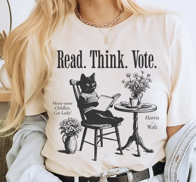 a beige tshirt with an illlustration of a black cat sitting at a table reading a book with text that says read. think vote